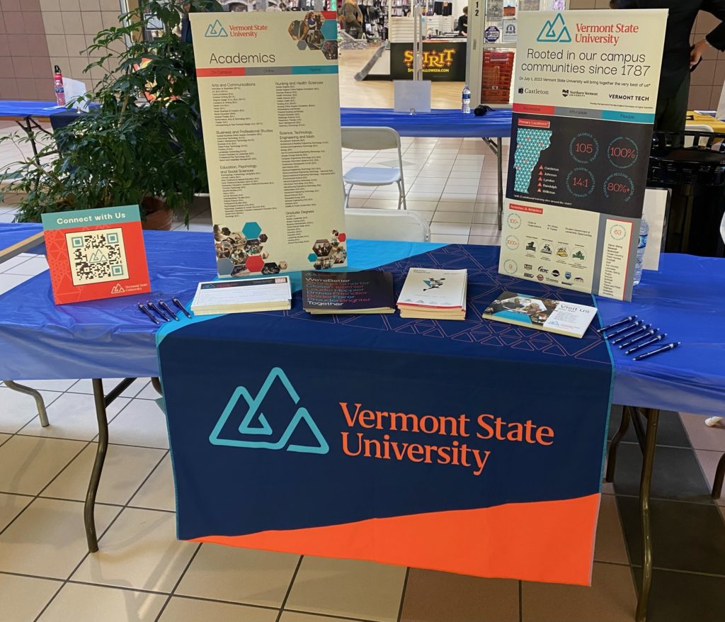 a picture of a table at a college recruiting event. It has a vermont state university banner, as well as flyers, pens, and two displays of vermont state university information and swag.