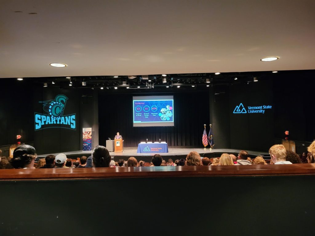 The image is taken from the back of a theater. it shows a room filled with prospective students, listening to what is being said. information about vermont state university is displayed on a screen, and the logo for vermont state and the Spartans logo are on either side of this screen